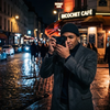 A man in a stylish coat holds a black Ricoh GR camera on a wet city street at night, surrounded by glowing café lights and cobblestone reflections.