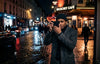 A man in a stylish coat holds a black Ricoh GR camera on a wet city street at night, surrounded by glowing café lights and cobblestone reflections.